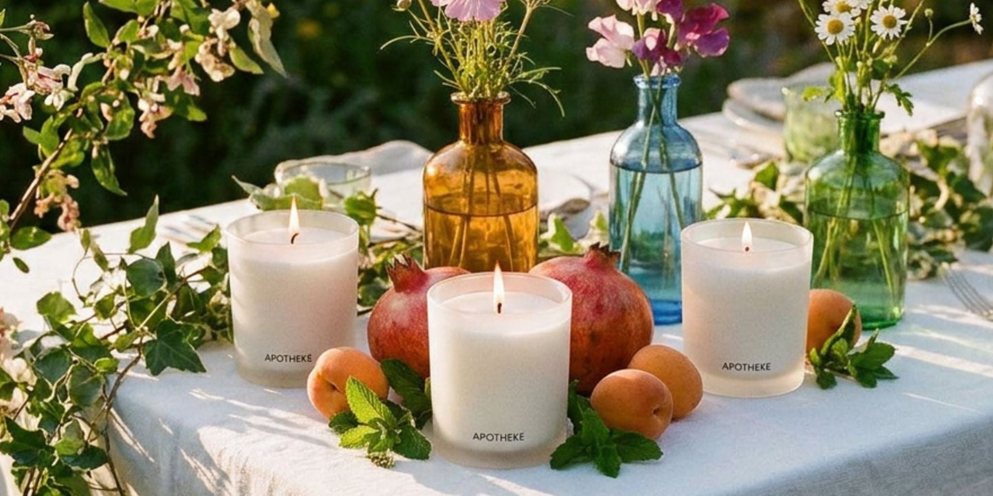 Three white pillar candles labeled 'APOTHEER' lit on a white tablecloth, styled with apricots, pomegranates, green leaves, and decorative glass vases in an outdoor garden setting